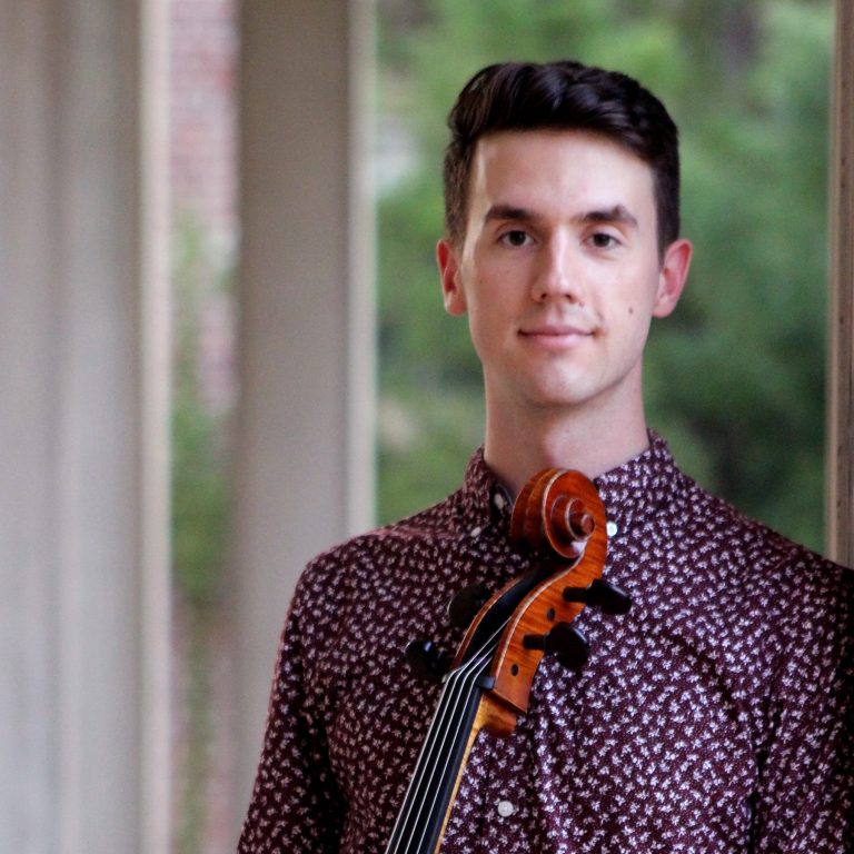 A young man with dark hair holds a violin, standing by a window in a patterned burgundy shirt, looking confidently at the camera.