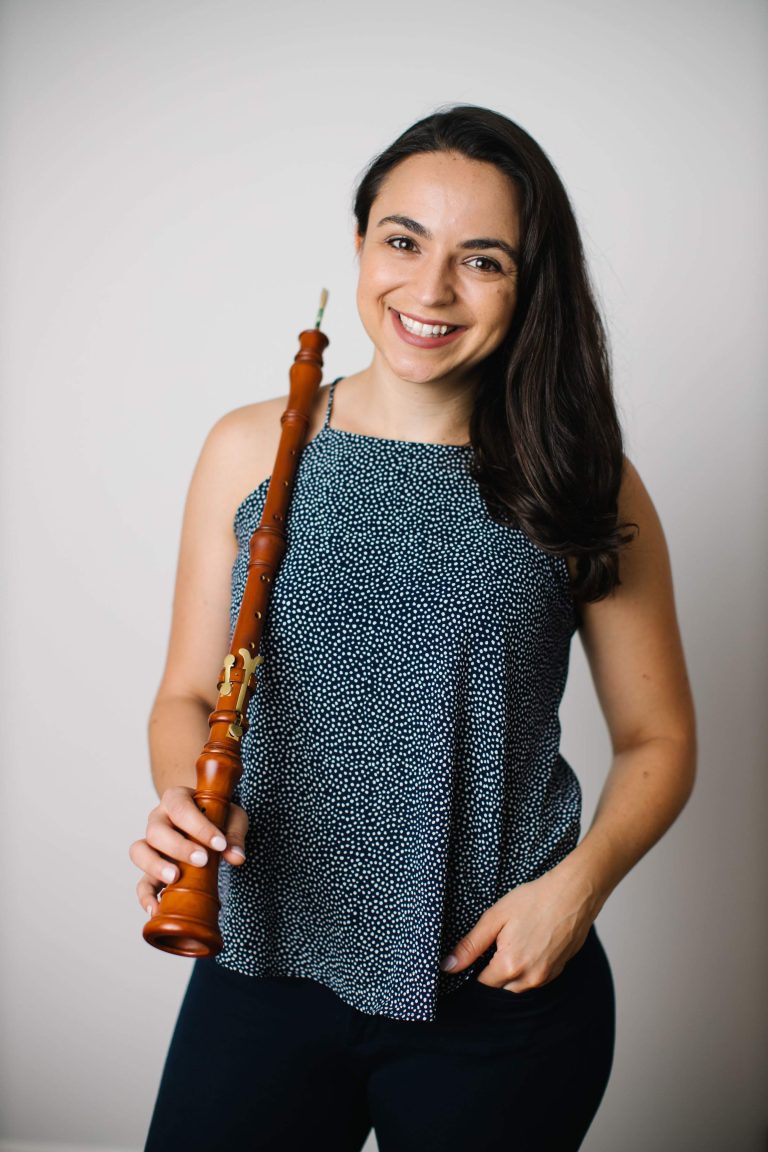 Smiling woman holding a wooden recorder, standing against a plain white background. she wears a sleeveless, dotted top and black pants.