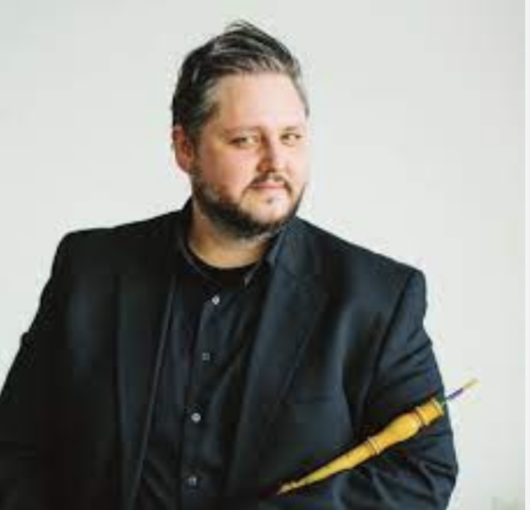 A man with a beard, dressed in a black shirt and blazer, holding a pencil and looking thoughtfully to the side.