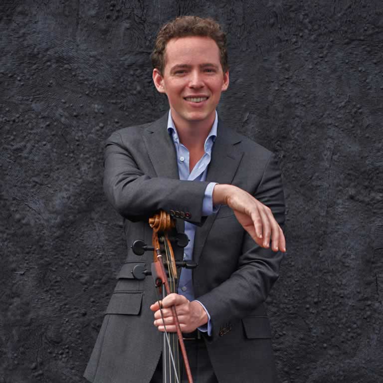 A man in a gray suit and blue shirt stands smiling, gesturing with his right hand, with a microphone stand in front of him against a black textured background.