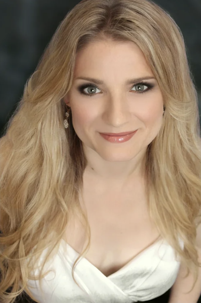 Portrait of a smiling woman with long blonde hair and a white dress, looking directly at the camera.