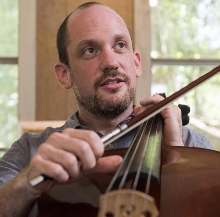 A man with a beard playing a cello, focusing intently on his instrument, indoors with natural light streaming in.