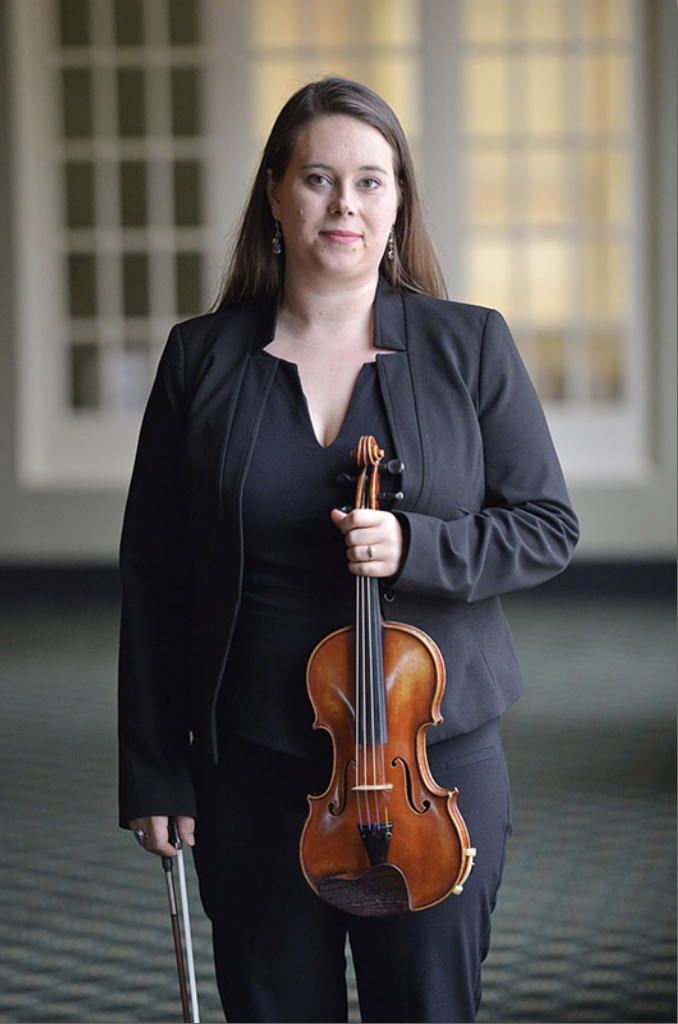 A woman in a black suit holds a violin and bow, standing confidently in a hallway with large windows and a patterned carpet.