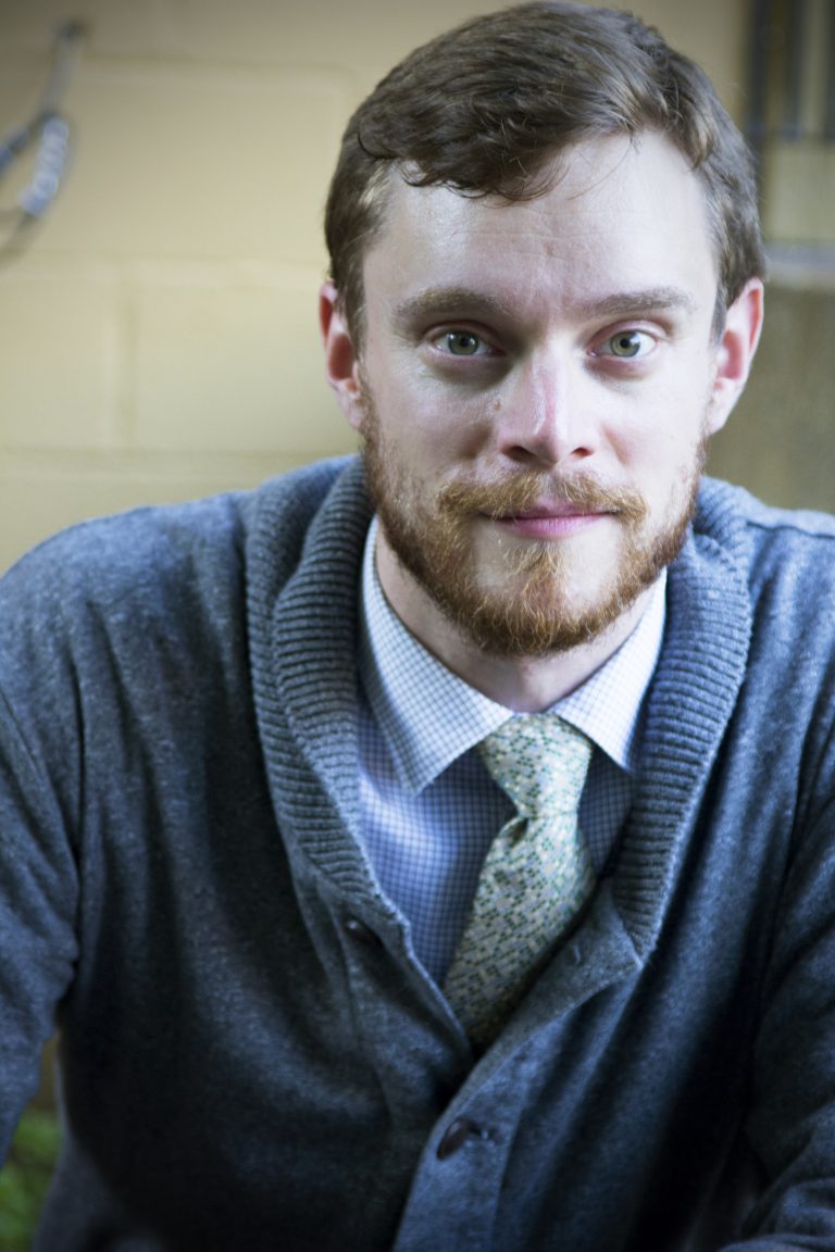 A young man with a beard and a slight smile, wearing a sweater over a collared shirt and tie, looks directly at the camera.