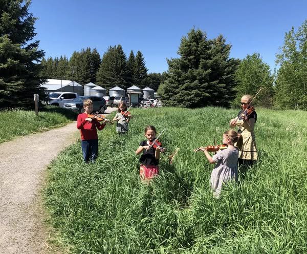 Five people, including children and an adult, play violins in a grassy field near trees, a path, parked cars, and farm silos under a clear blue sky.