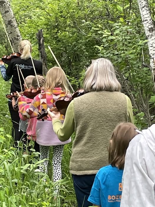 A group of people, including children and adults, walk through a green forest carrying violins on their shoulders.