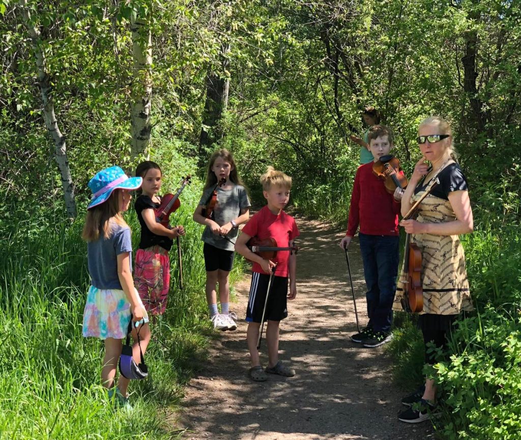 A group of children hold violins on a forest path with a woman holding a violin standing nearby, offering guidance. Trees and greenery surround them.