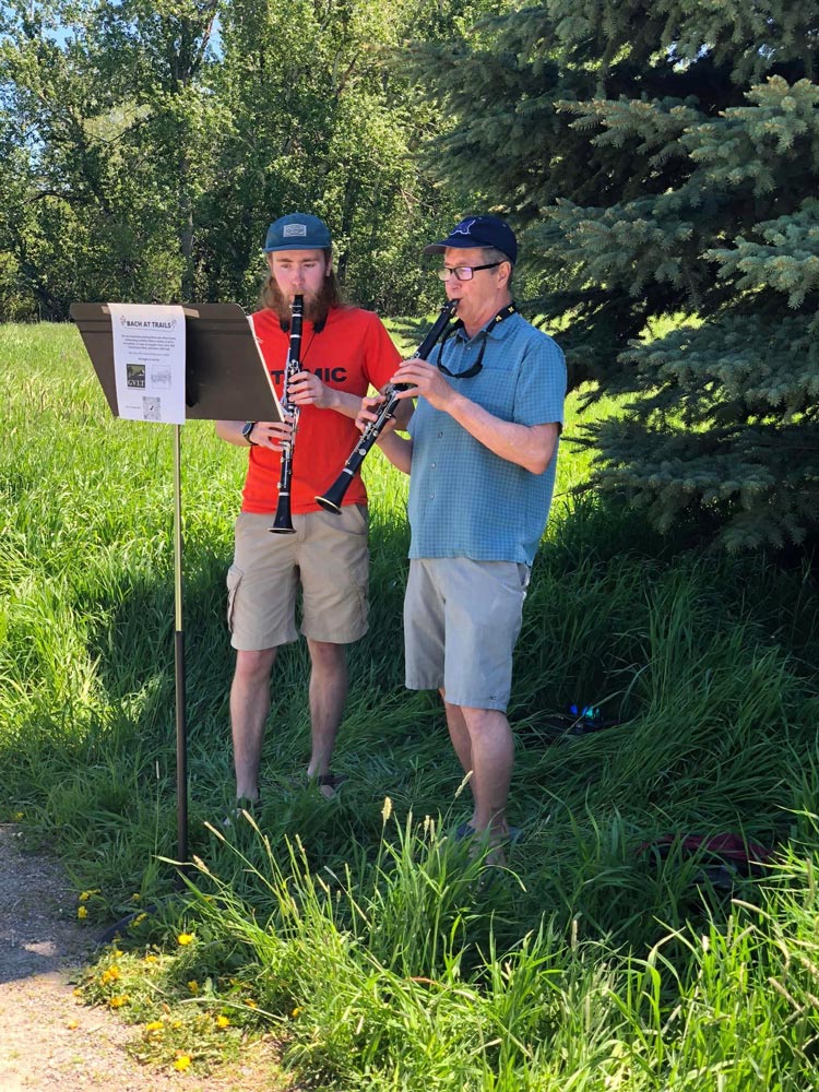 Two men wearing casual clothes and hats play clarinets outside next to a music stand. They are standing on a grassy area with trees in the background.