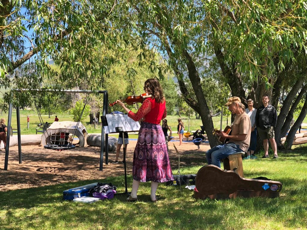A woman in a colorful dress plays violin while a man seated beside her plays a box drum in a park. A small audience watches, and children play on playground equipment in the background.