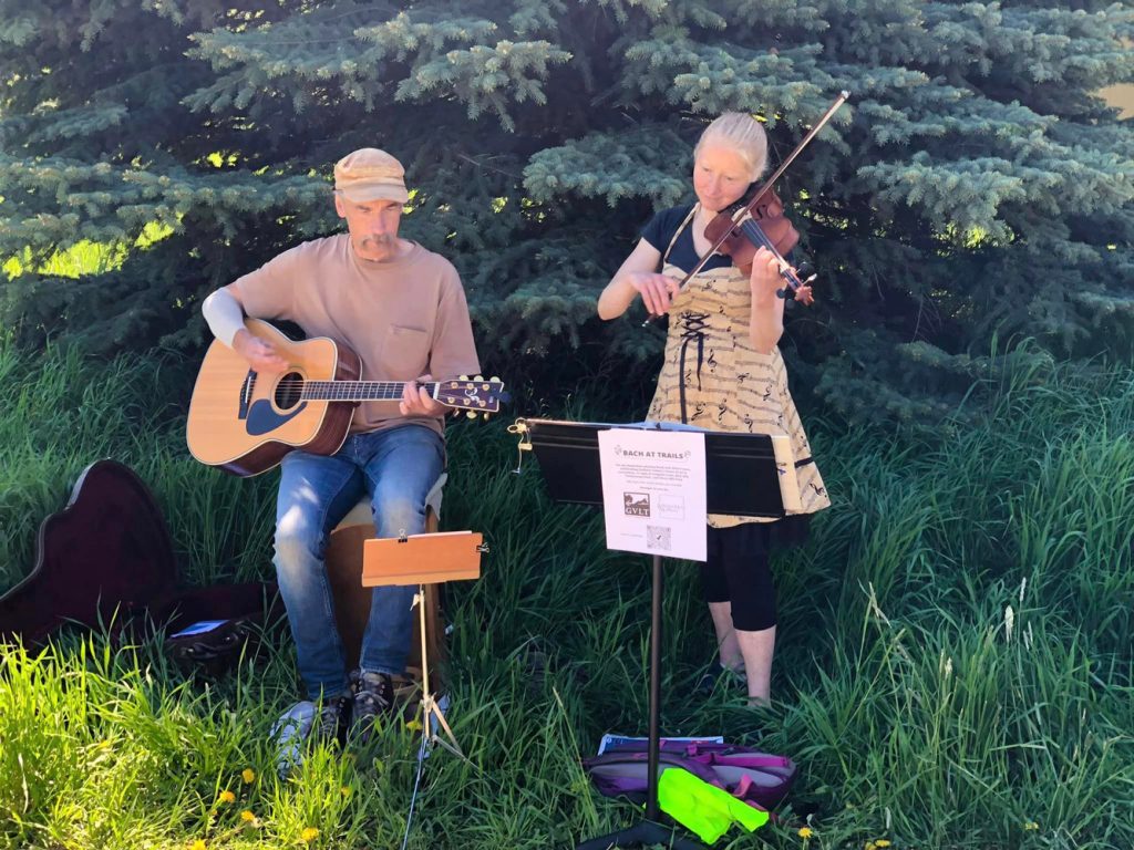 A man plays an acoustic guitar and a woman plays a violin outdoors near a pine tree. They have a music stand with sheet music in front of them and are surrounded by grass.