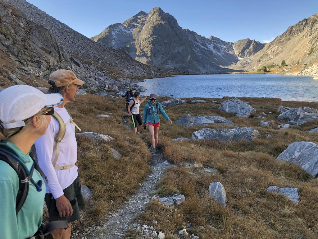A group of hikers follows a trail beside a mountain lake, surrounded by rocky peaks and grassy terrain.