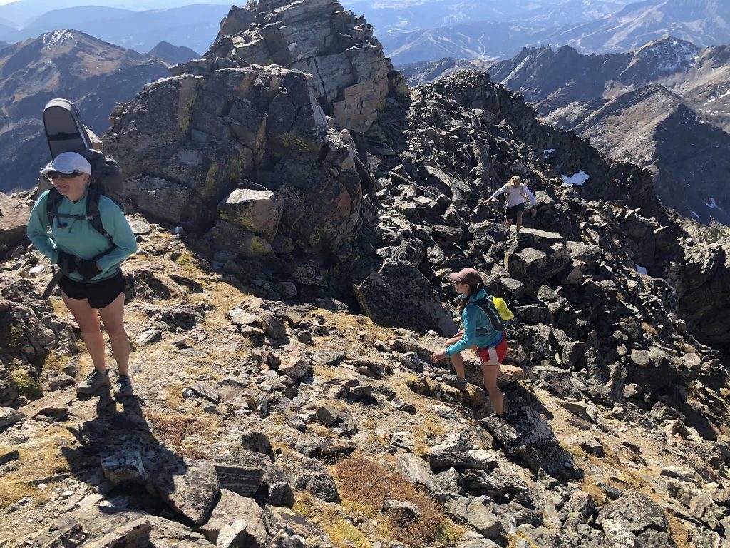 Three hikers navigate a rocky mountain ridge under a clear sky, with distant peaks in the background.