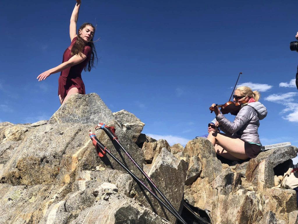 A woman dancing on rocks while another woman plays a violin. Ski poles are resting nearby under a clear blue sky.