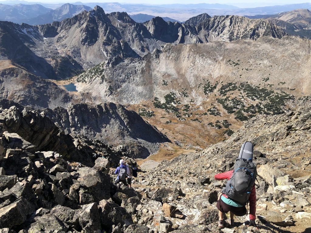 Hikers descend a rocky mountain trail with a panoramic view of peaks and valleys under a clear sky.