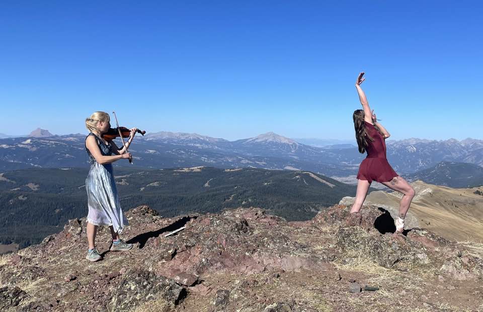 Two women perform on a rocky mountain peak: one plays a violin, and the other dances gracefully. The background displays a vast landscape and blue sky.