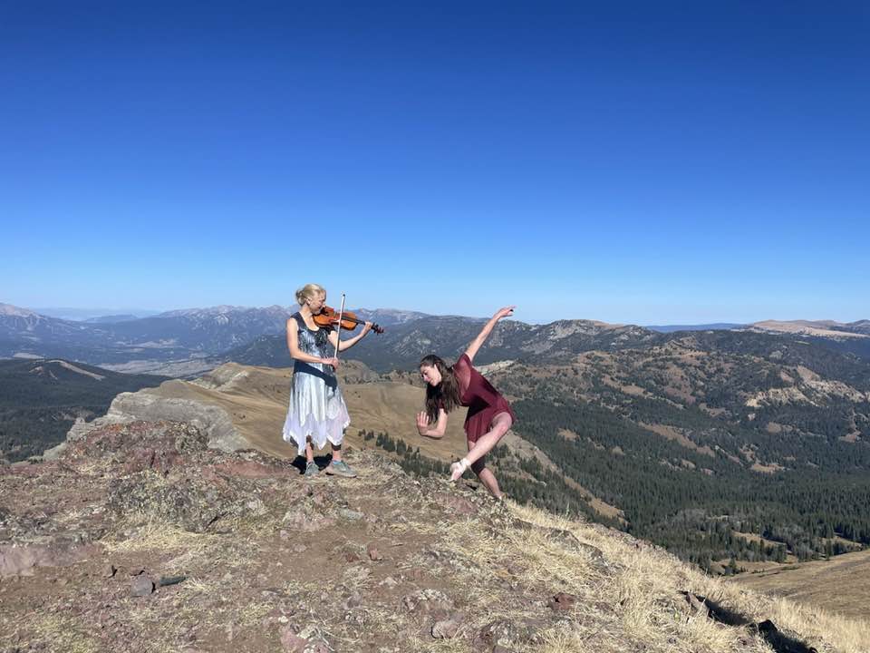 Two women stand on a mountain peak, one playing a violin and the other dancing, with a vast landscape and clear blue sky in the background.