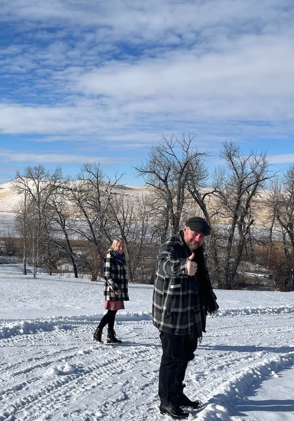 Two people in plaid jackets stand on a snowy path with bare trees and hills in the background. One person gives a thumbs-up.