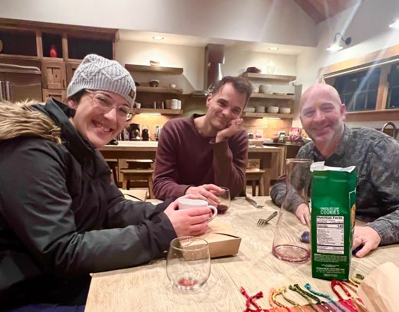Three people sitting at a wooden table in a kitchen. They are smiling and have drinks. The kitchen has wooden cabinetry and shelves with plates. A bag of cookies is on the table.