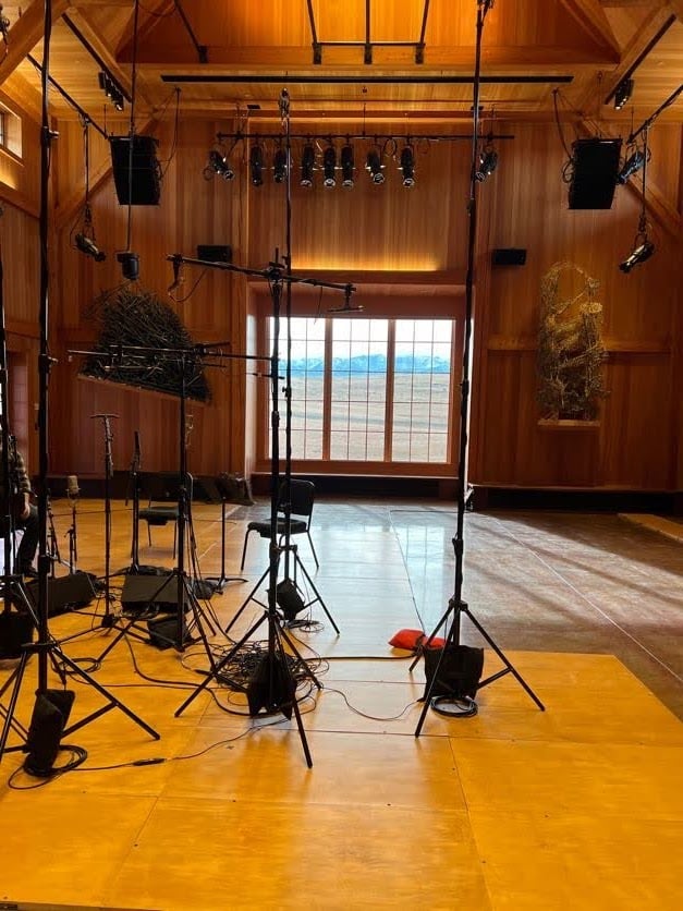 Recording studio with wooden walls, large window, microphones, and stands on a wooden floor. Stage lighting is visible above.