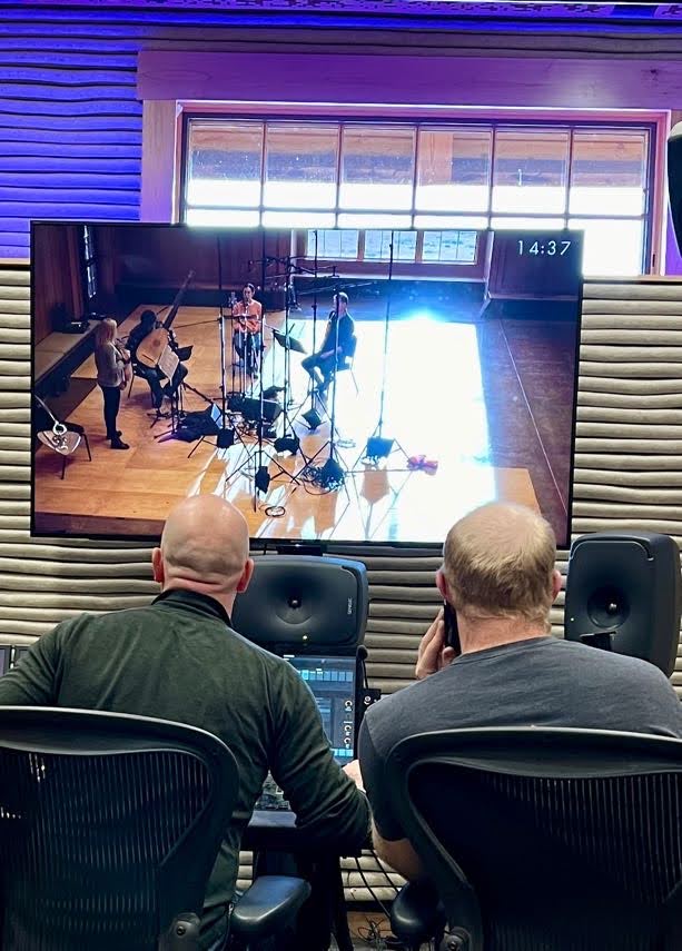 Two men in a recording studio observe musicians performing on a large screen, with sound equipment in the foreground.