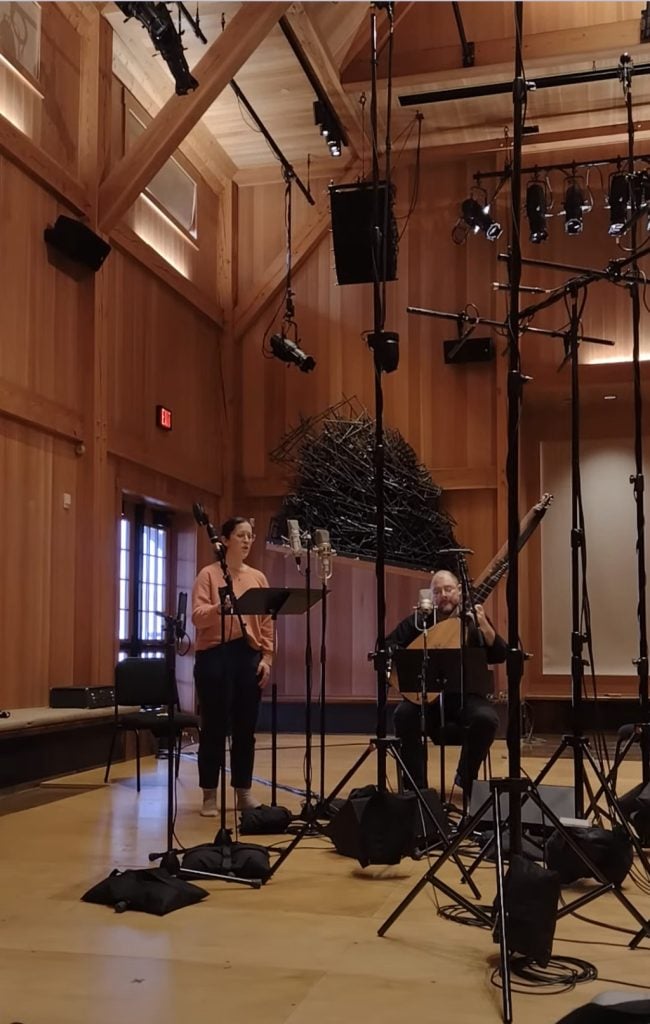 A woman stands singing into a microphone, while a man seated next to her plays a lute in a wooden recording studio surrounded by microphones and audio equipment.