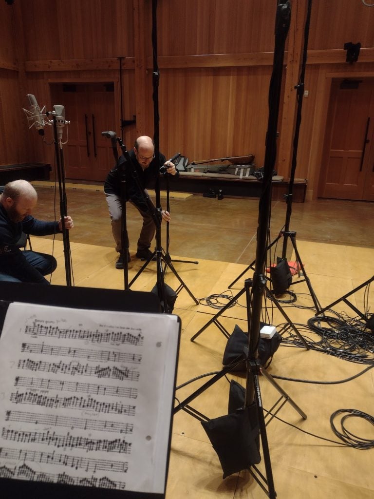 Two people adjust microphone stands in a recording studio with sheet music visible in the foreground.