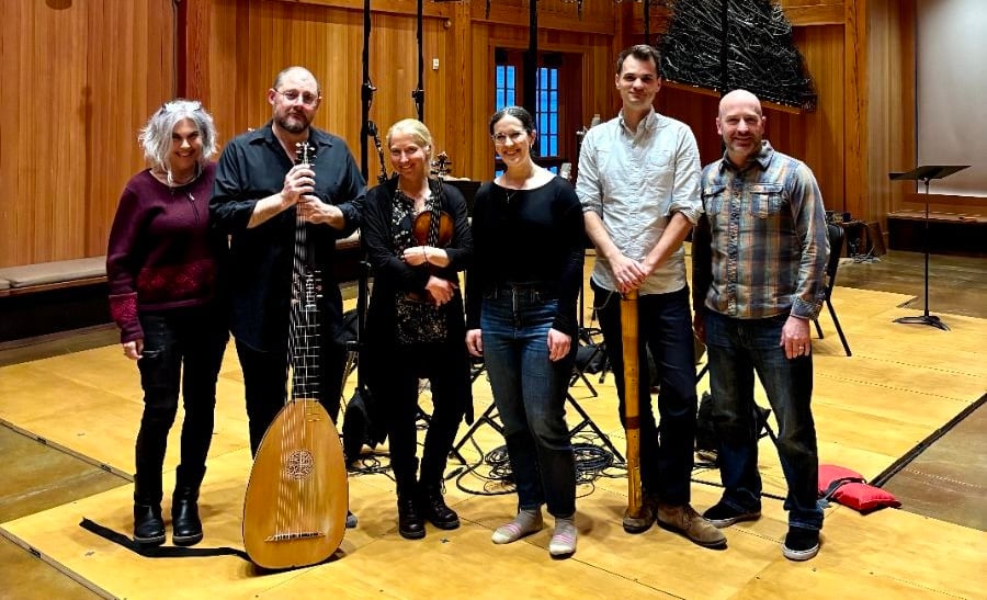 A group of six people stand together in a wooden-floored room with recording equipment. One person holds a lute-like instrument.