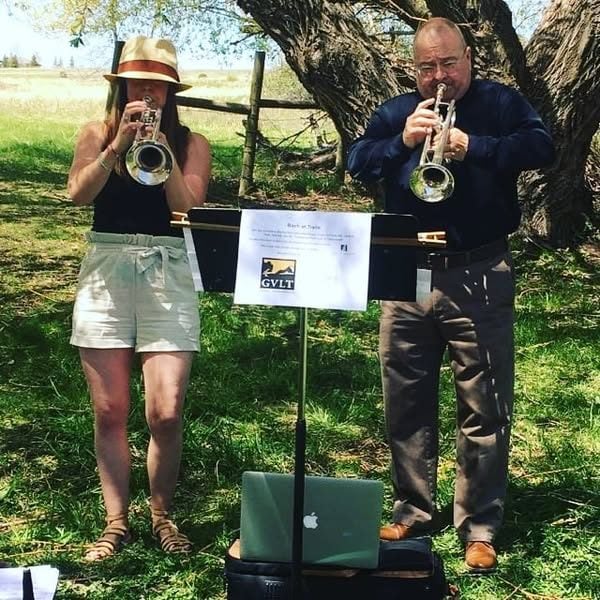 A woman and a man play trumpets outdoors under a tree, standing behind music stands and a laptop, with sunlight and grass around them.