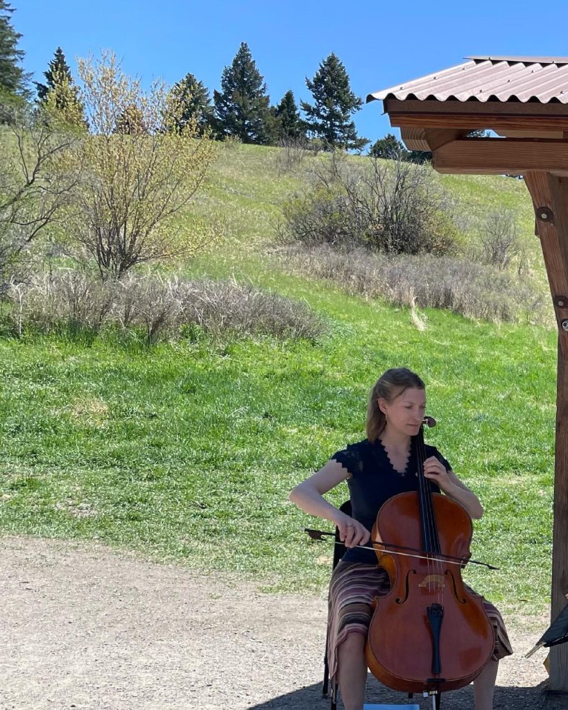 A woman sits outdoors on a sunny day, playing a cello near a wooden structure with grassy hills and trees in the background.