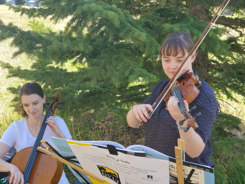 Two women play string instruments outdoors; one is playing a cello, the other a violin, with sheet music clipped to stands in front of them and trees in the background.