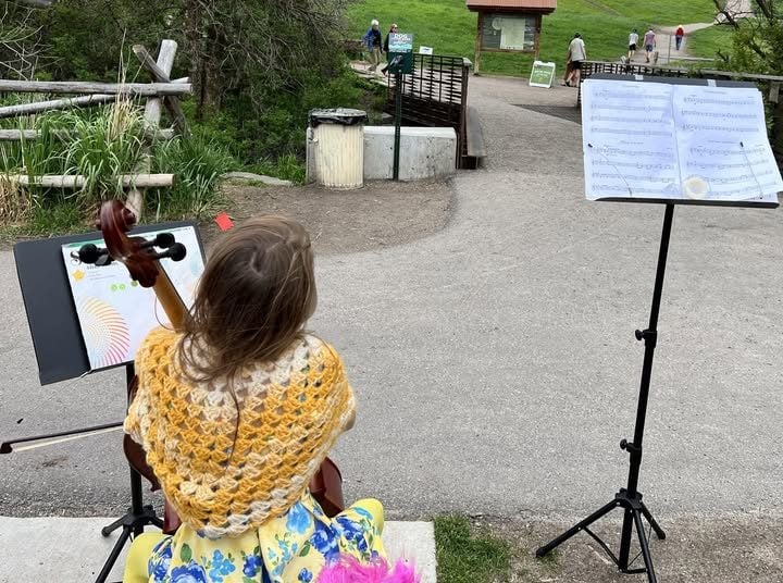 A child sits outdoors playing a cello, with sheet music on a stand in front of her, near a path and a small bridge in a park setting.