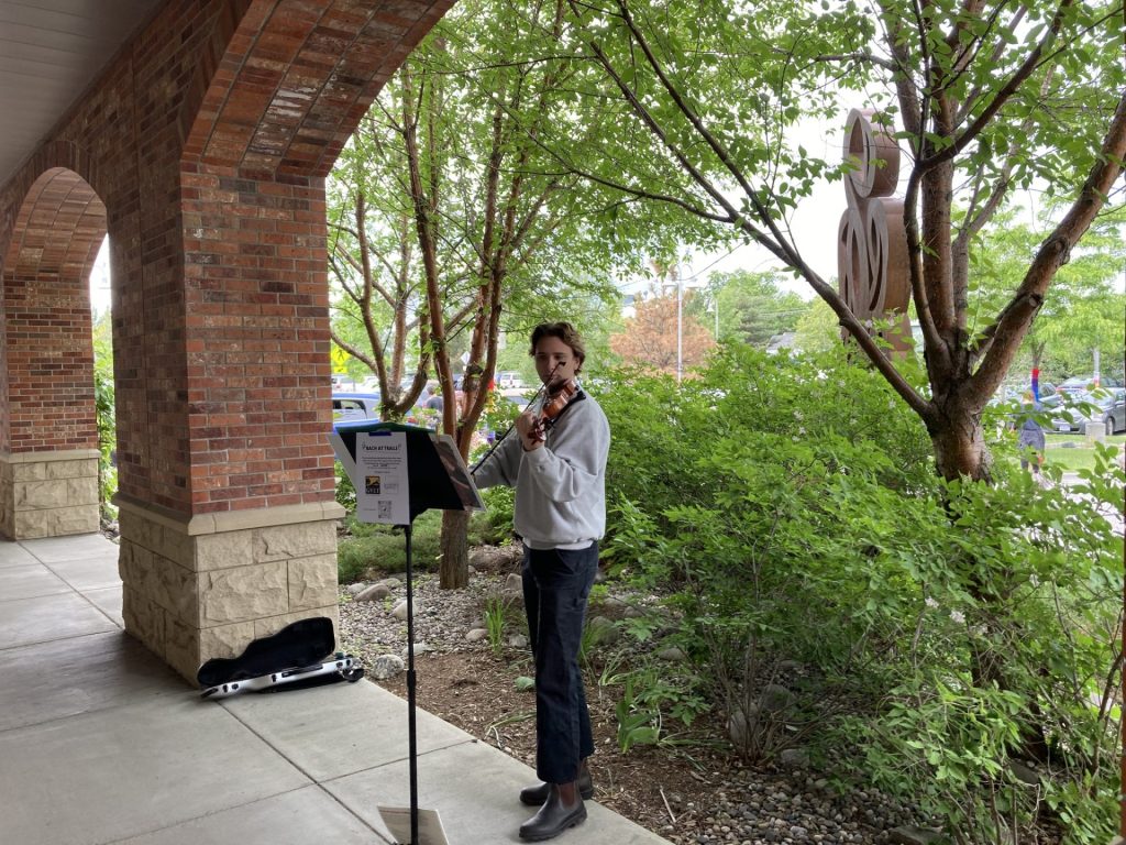 A person plays violin outside under an archway near a brick building, standing beside a music stand, with greenery and parked cars in the background.