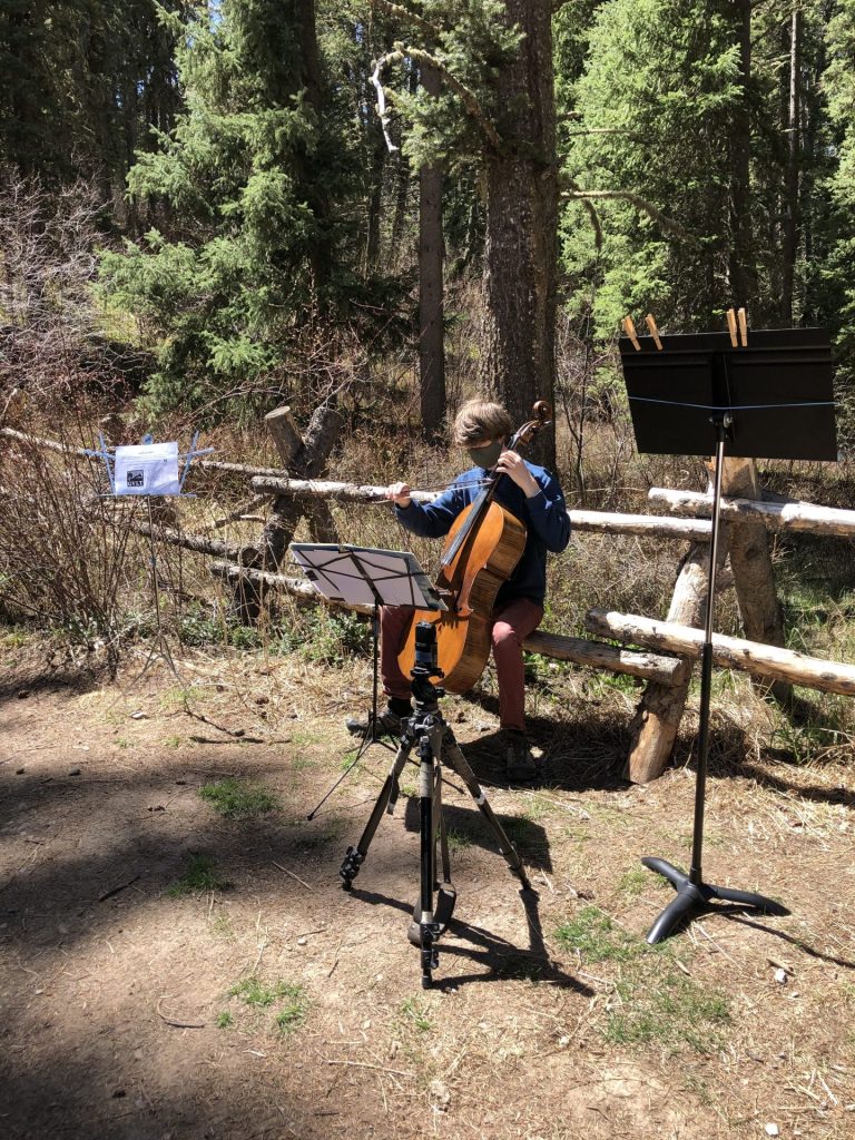 A person plays a cello outdoors in a forest, seated near a wooden fence, with sheet music attached to a stand and a tripod set up nearby.