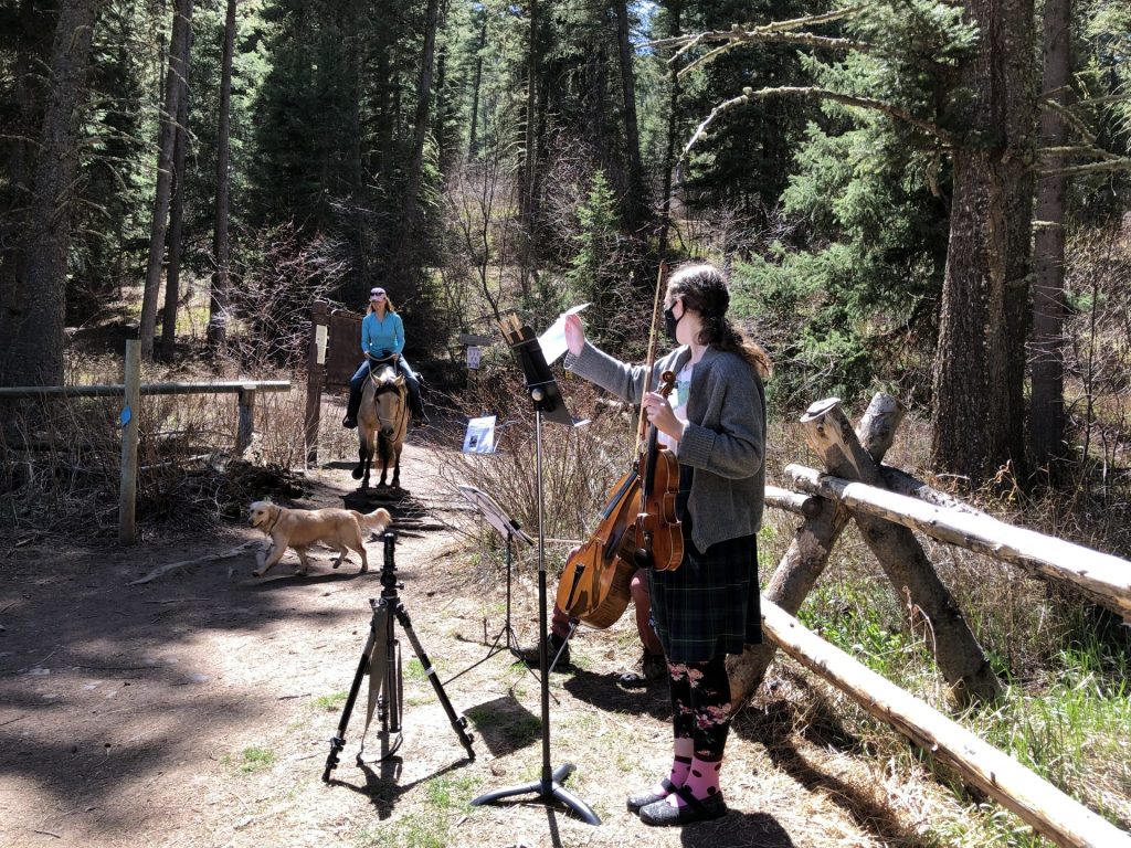 A person plays cello with a microphone setup on a forest trail while a horse rider and two dogs pass by in the background.