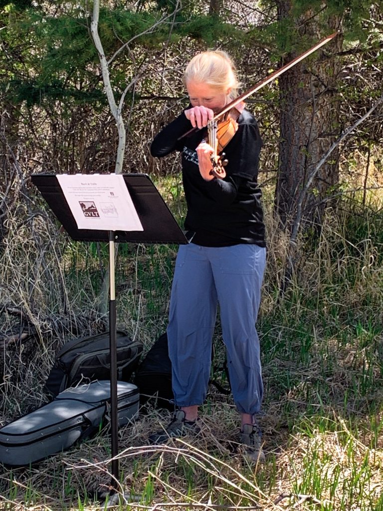 A person plays a violin outdoors in a wooded area, standing next to a music stand with sheet music and an open violin case on the ground.