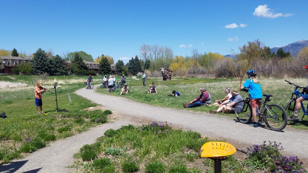 A group of people, some on bikes and others sitting on grass, watch a child speaking into a microphone along a park path on a sunny day.