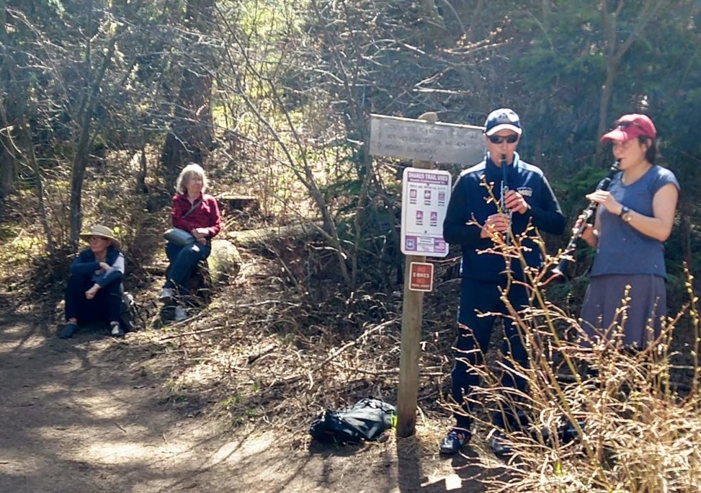Two people play clarinets next to a trail sign in a wooded area while two others sit nearby on the ground and a log.