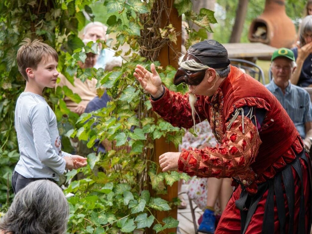 A performer in a Venetian-style mask and ornate costume interacts with a smiling child outdoors as onlookers watch.