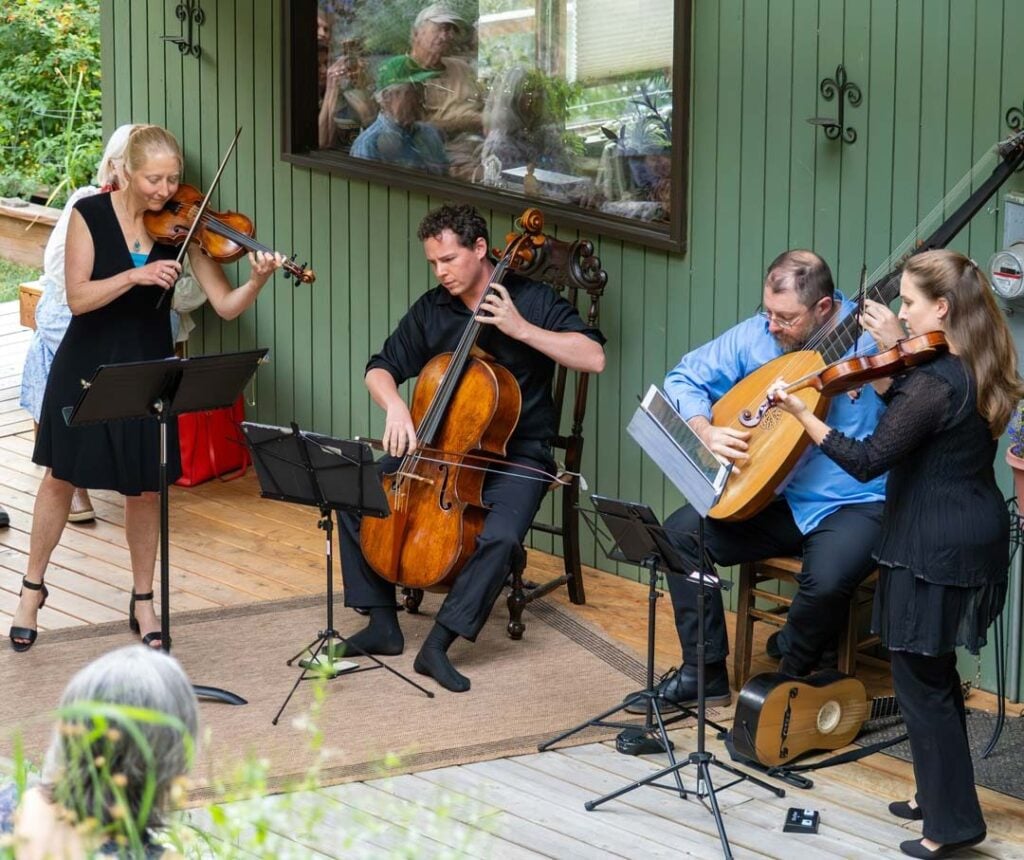 Four musicians perform with a violin, cello, lute, and viola on a wooden deck beside a green building, with an audience seated nearby.