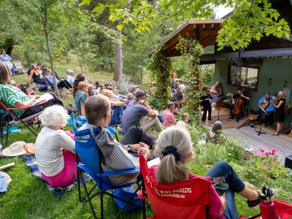 An audience sits on a grassy slope watching a small string ensemble perform on an outdoor stage surrounded by trees and greenery.