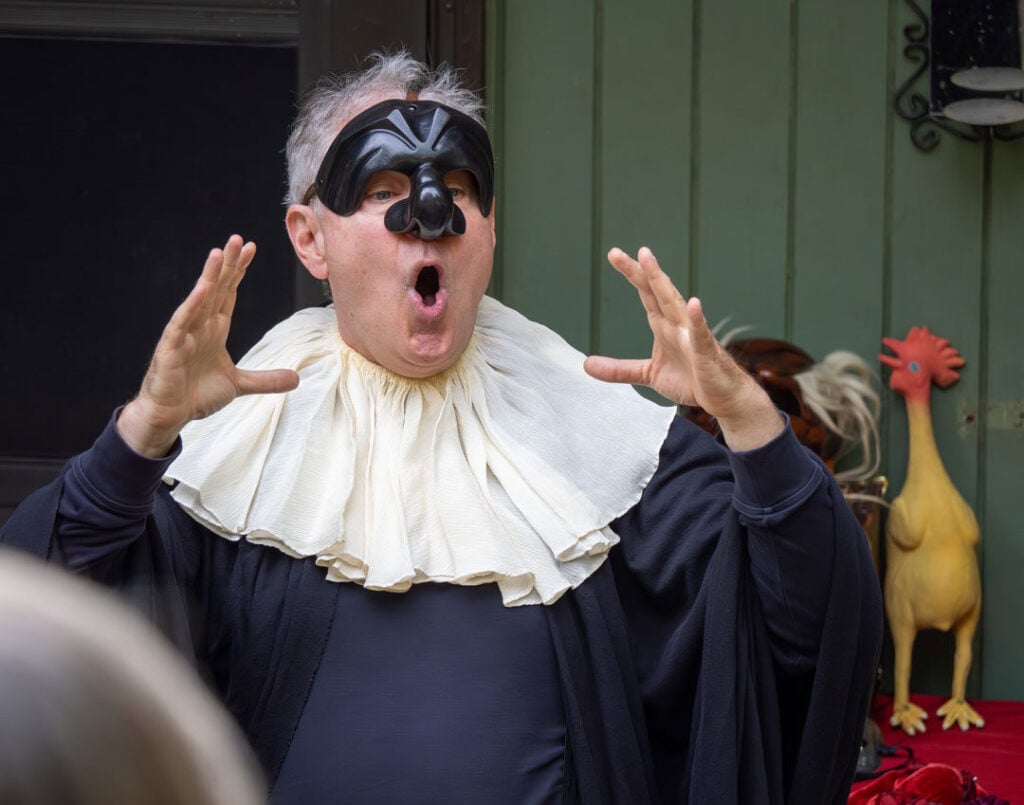 A person wearing a theatrical mask and large white ruffled collar gestures dramatically with open mouth; a rubber chicken is visible in the background.