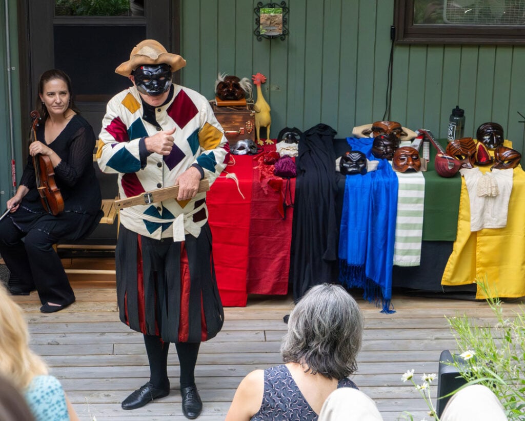 A performer in a colorful costume and mask stands on a porch stage, gesturing, with masks and props displayed on tables behind him; an audience and musician are present.
