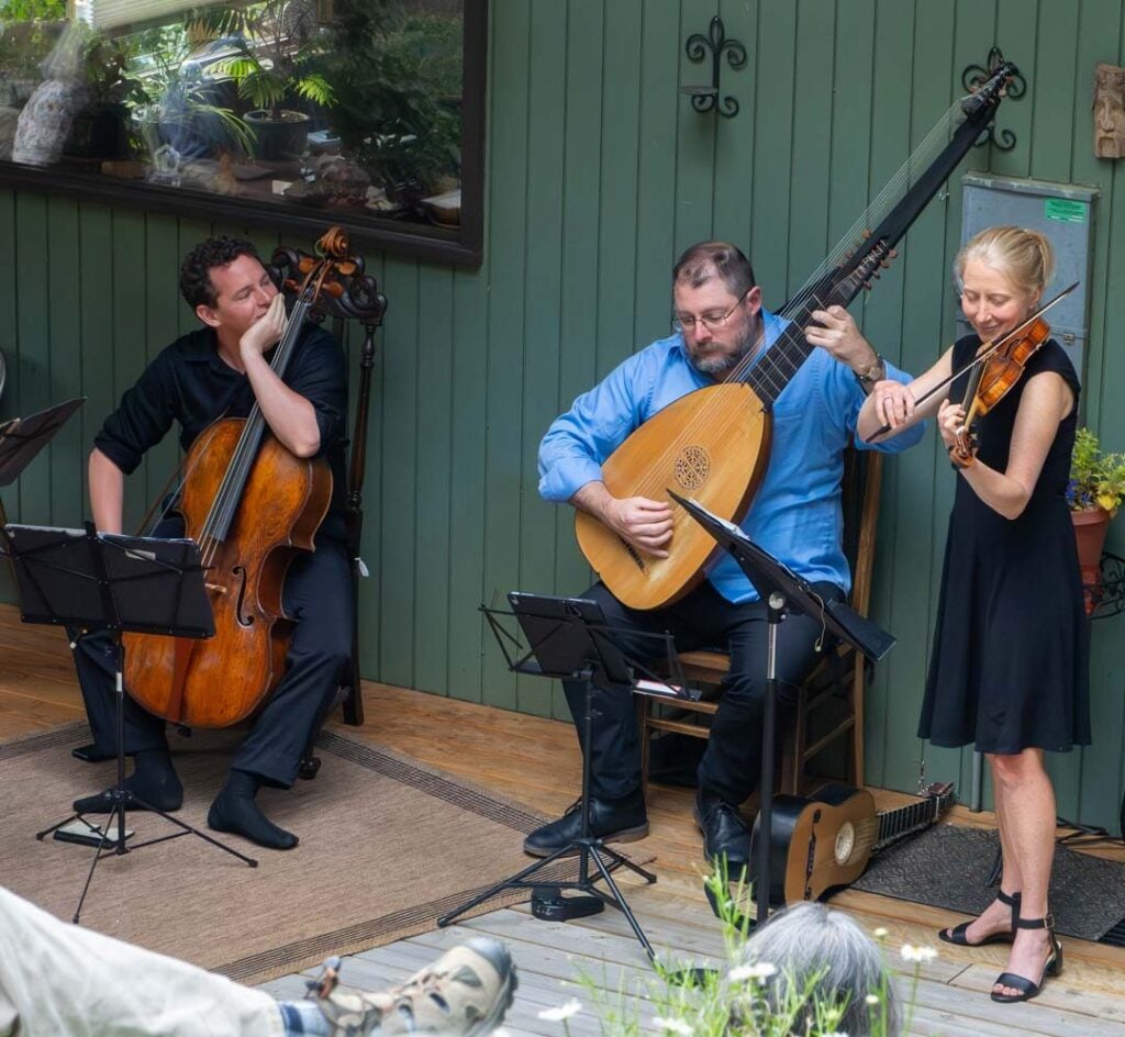 Three musicians perform together: one playing a cello, one playing a lute, and one playing a violin, seated and standing on a porch with sheet music stands in front of them.