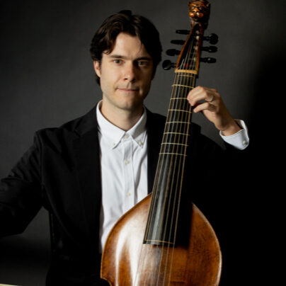 A man in a suit plays a large stringed instrument with a dark background.