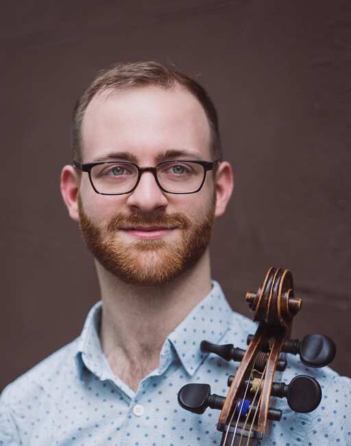 A man with glasses and a beard, wearing a light blue patterned shirt, stands in front of a brown background with a cello resting on his shoulder.